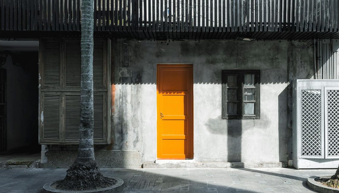 Vibrant orange door contrasts with a rustic concrete wall adorned with windows.