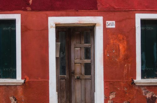 Vibrant red and orange facade of a Venetian house featuring a rustic wooden door and paved sidewalk.