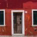 Vibrant red and orange facade of a Venetian house featuring a rustic wooden door and paved sidewalk.