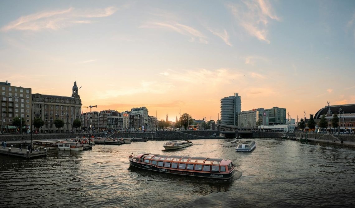 Scenic view of Amsterdam canals at sunset with tour boats cruising and cityscape backdrop.
