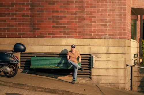 A person sits on a bench beside a brick building wall, with a parked scooter nearby, embodying urban relaxation.