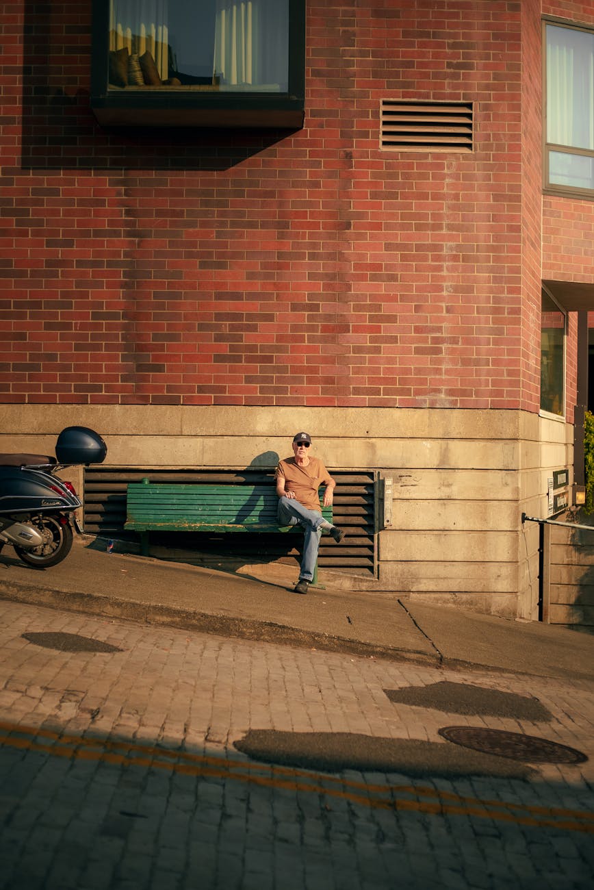 A person sits on a bench beside a brick building wall, with a parked scooter nearby, embodying urban relaxation.