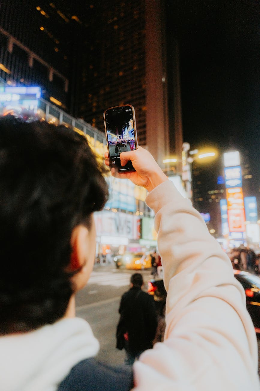 Tourist photographing vibrant city streets at night with a smartphone, capturing urban energy.