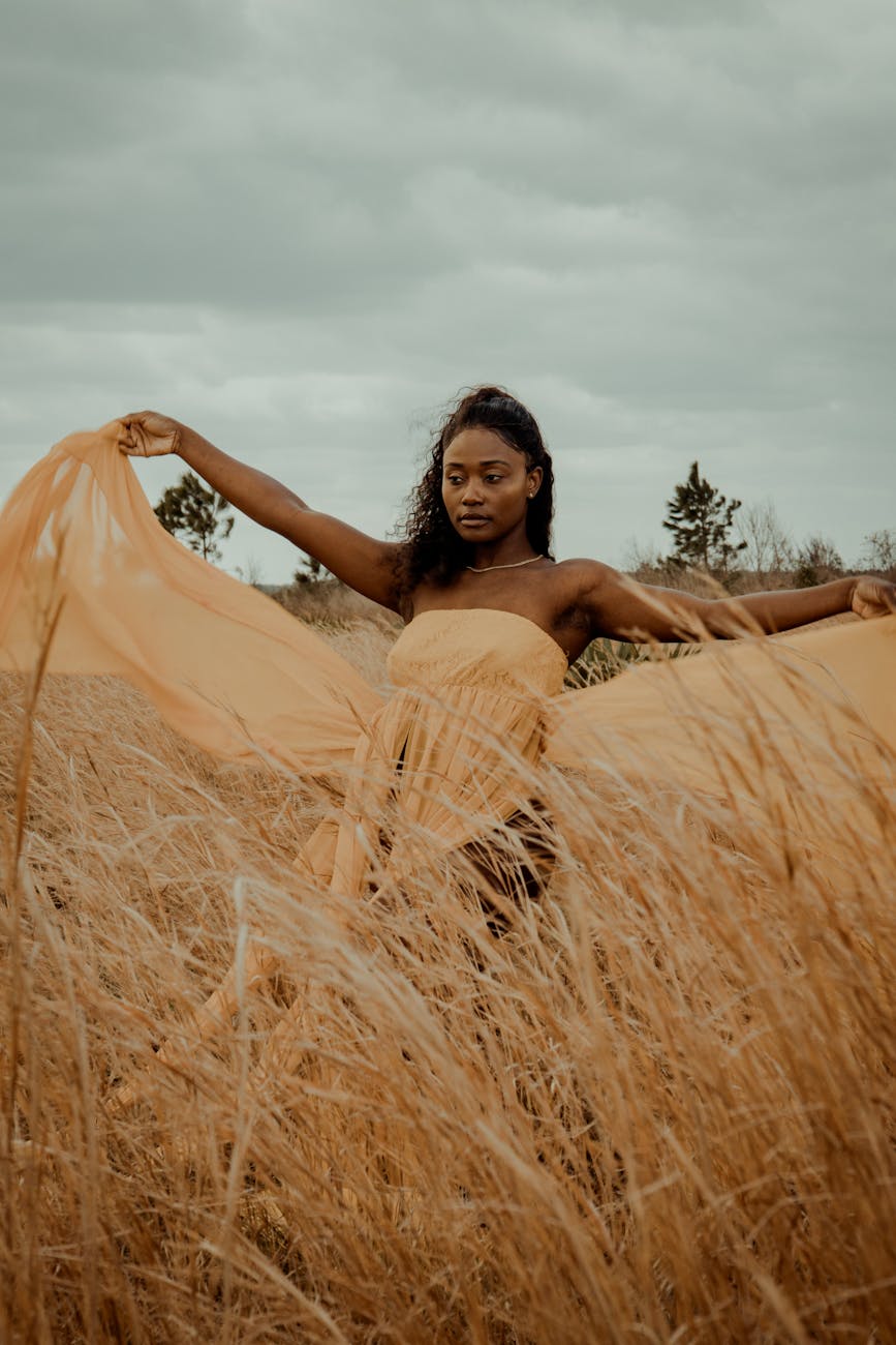 Graceful woman in flowing dress embraced by the golden wheat field under a cloudy sky.